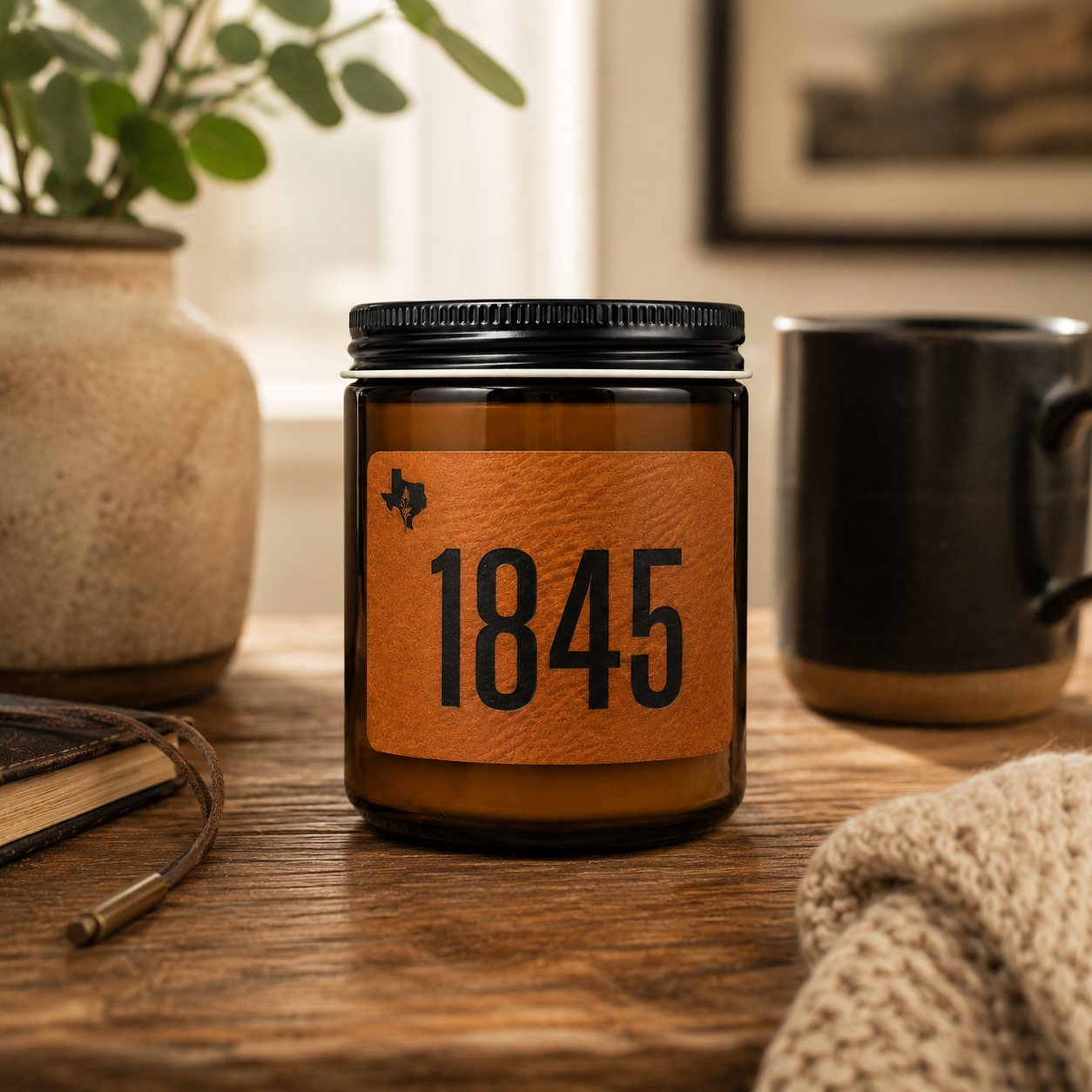 Candle jar with '1845' label on a wooden surface with a mug and plant in the background