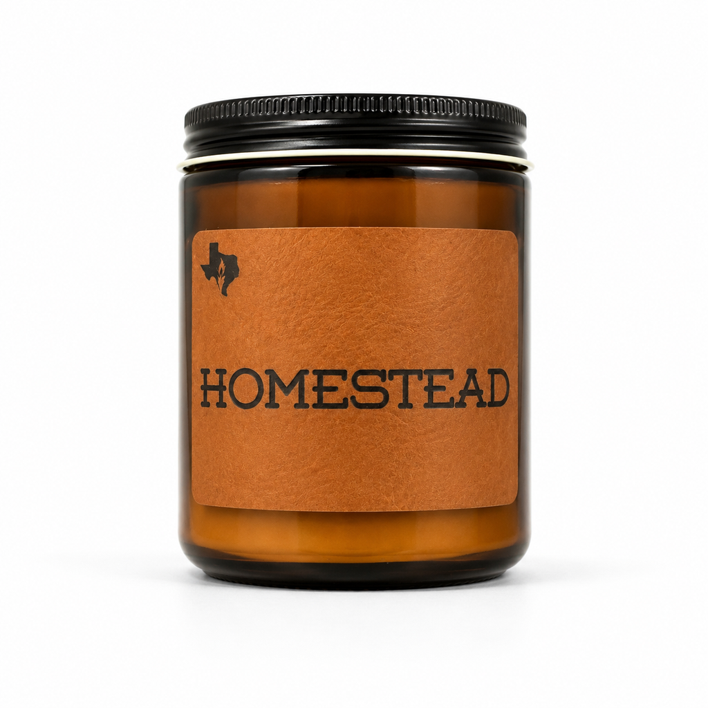 The Homestead 8oz soy candle in an amber glass jar with a black lid and a leather label reading 'Homestead', hand-poured by Texas Candles.