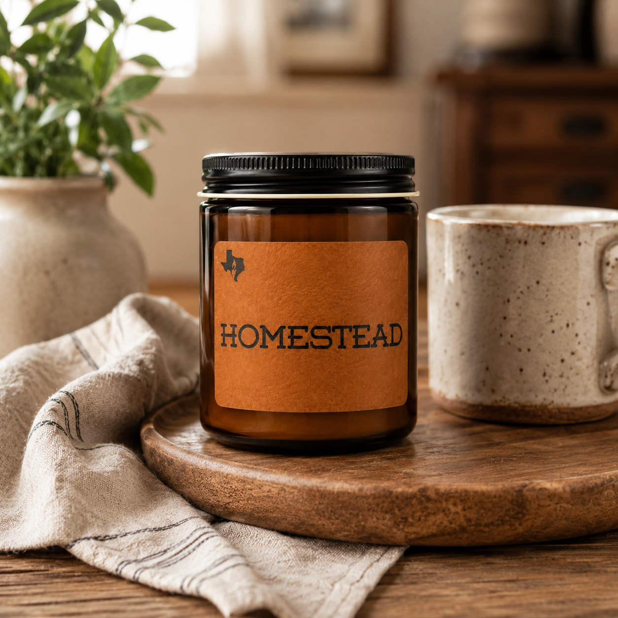 The Homestead 8oz candle on a round wooden board with a ceramic vase of greenery, a speckled mug, and a linen napkin.