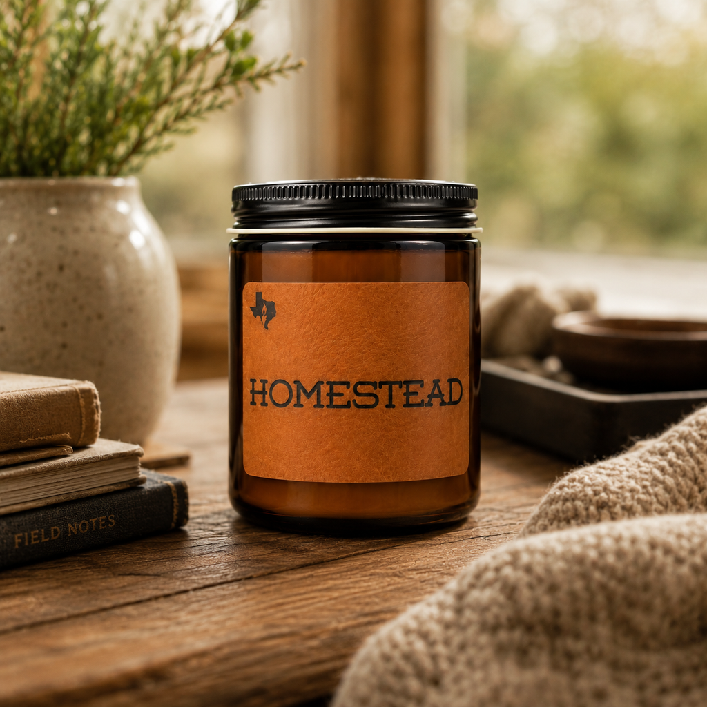 The Homestead 8oz candle on a wooden table by a window with a vase of greenery, stacked books, and a knit throw.