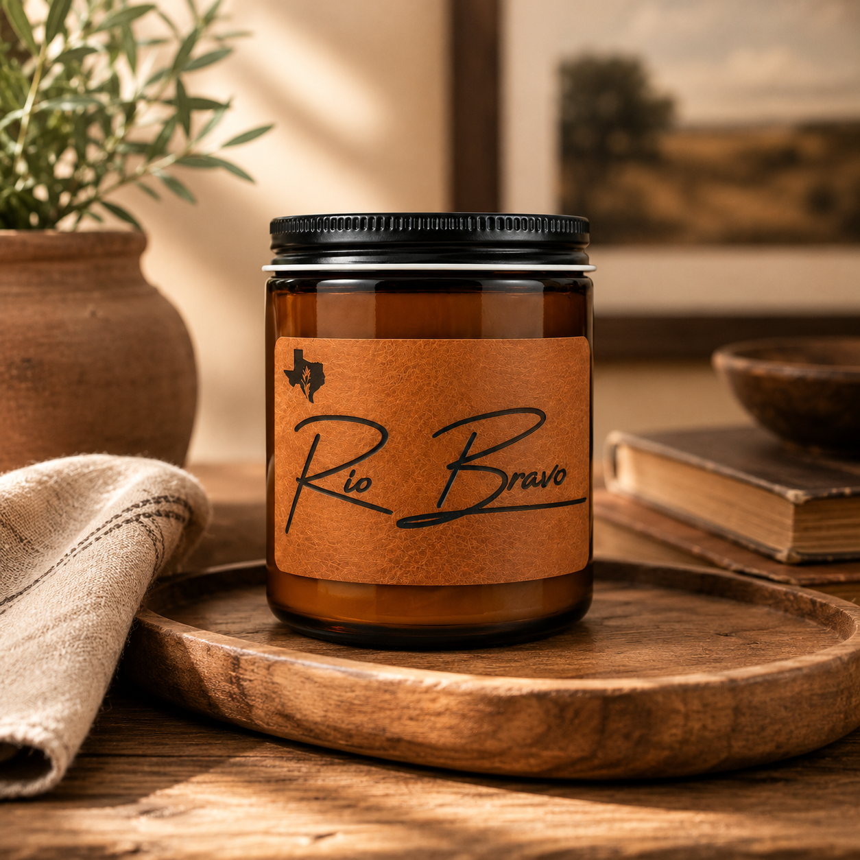 Rio Bravo 8oz candle on a wooden tray beside a terracotta pot of greenery and a worn book, with a framed landscape behind.